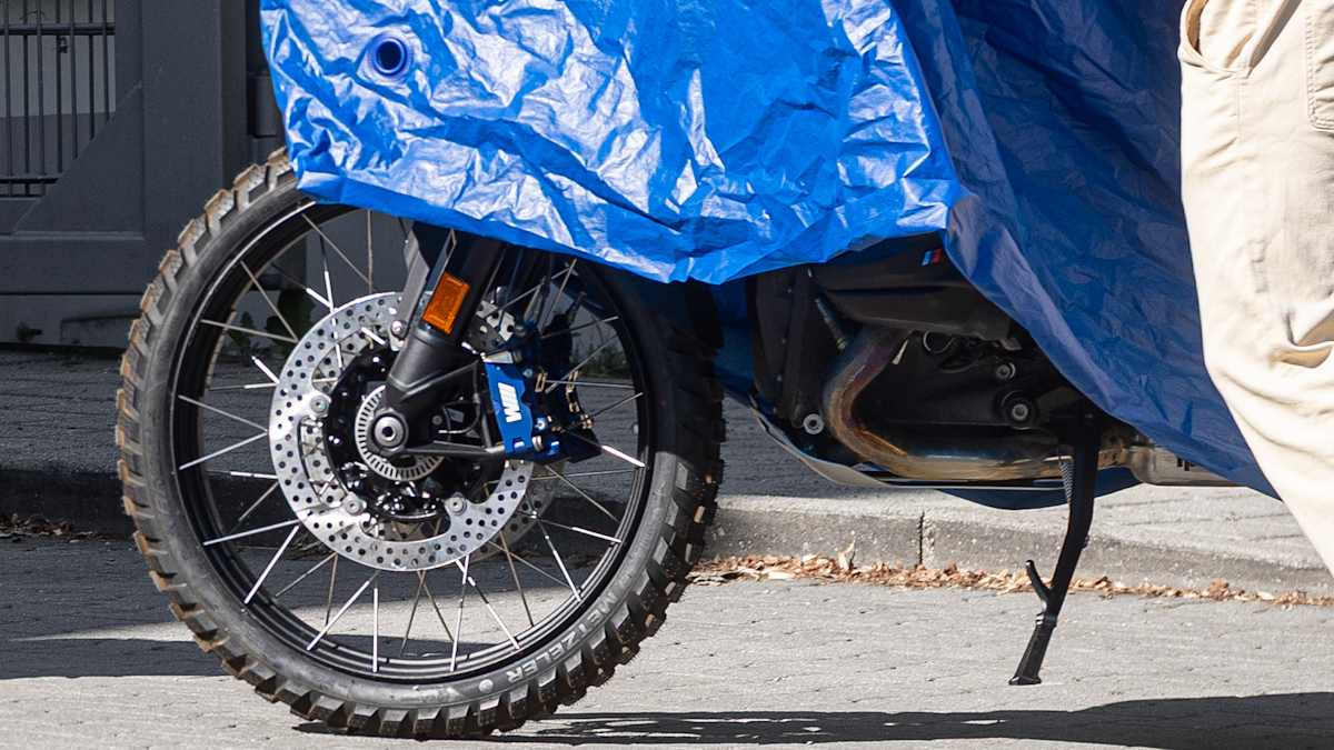 Close-up of the BMW M1300GS prototype's front wheel, showing the blue M-branded brake caliper and Metzeler Karoo off-road tire under a blue tarp