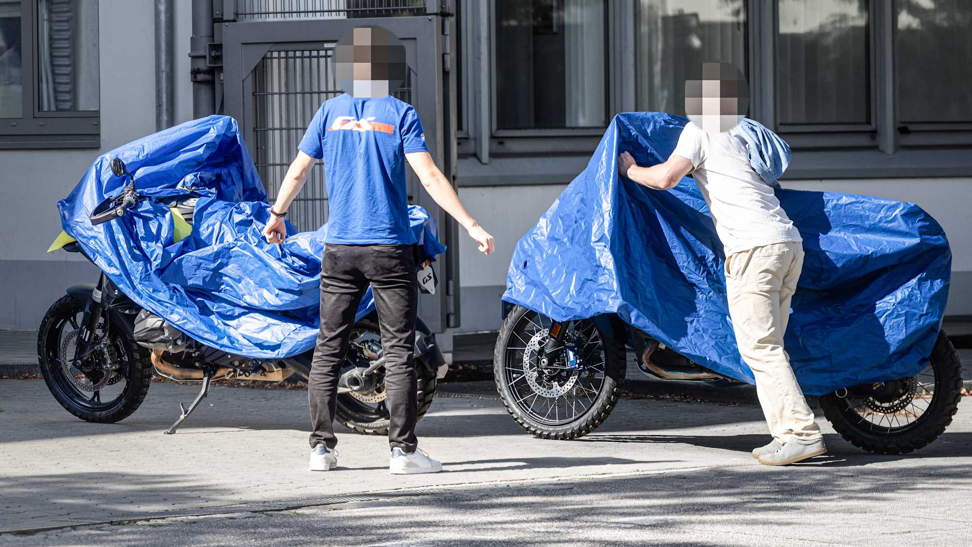 Two camouflaged BMW GS prototypes under blue tarps with spoked wheels and Metzeler Karoo off-road tires visible, parked side by side outside a BMW facility