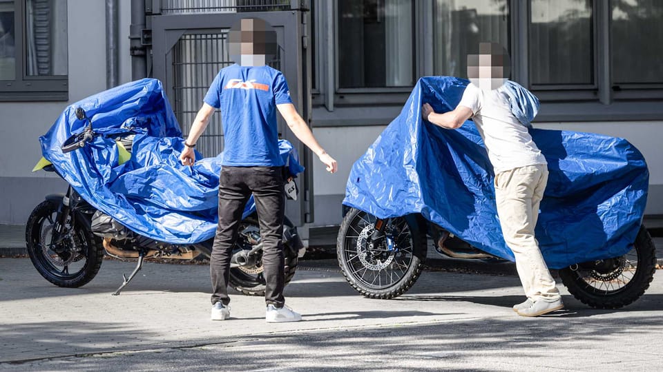 Two camouflaged BMW GS prototypes under blue tarps with spoked wheels and Metzeler Karoo off-road tires visible, parked side by side outside a BMW facility